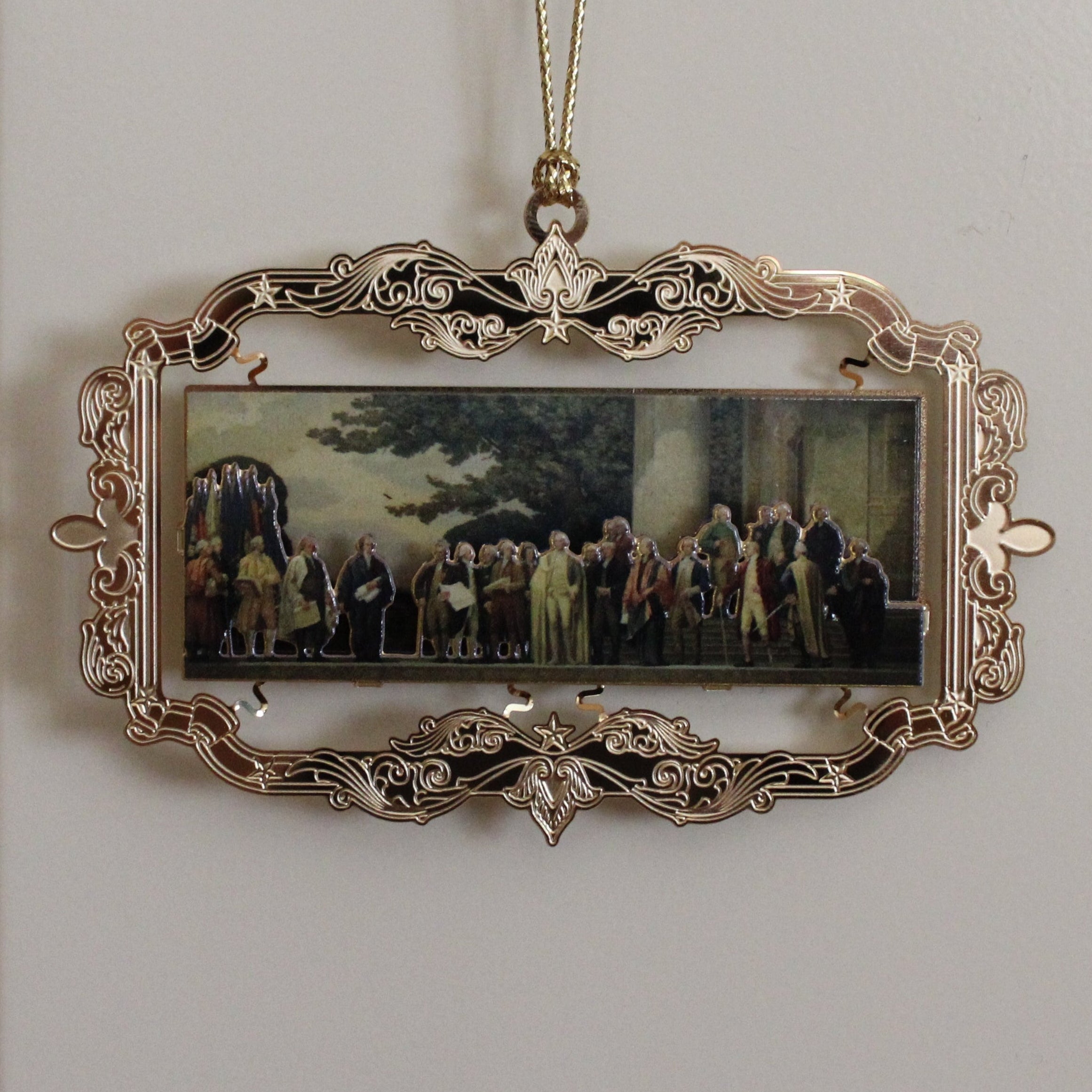 Decorative ornament with a miniature mural painting of a group of Founding Fathers in a gold-colored frame, hanging on a white wall.