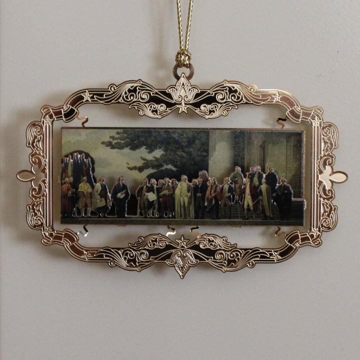 Decorative ornament with a miniature mural painting of a group of Founding Fathers in a gold-colored frame, hanging on a white wall.