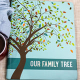 Our Family Tree Book: A graceful tree adorns the cover, with leaves in shades of spring green, olive, teal, orange, brick red, and gold foil. The words 'Our Family Tree' appear in white letters traced with gold foil. Smooth matte hardcover with a subtle blue/green check pattern. Raised embossing lends texture and dimension. Rounded corners for reduced wear.